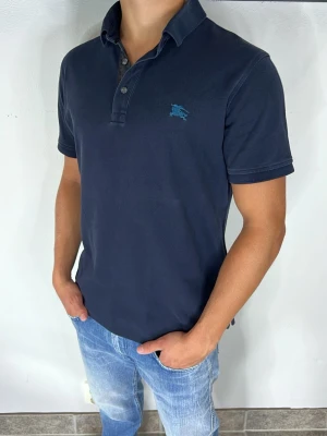 Burberry Piké - Snygg mörkblå pikétröja med klassisk krage och korta ärmar. Tröjan har knappar framtill och en liten broderad logga på bröstet. Perfekt till jeans för en avslappnad men stilren look.