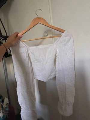 Vit topp med spetsdetaljer - Säljer en söt vit  topp med långa ärmar och broderade spetsdetaljer. Toppen har en croppad passform och elastiska muddar vid ärmsluten. Perfekt för sommarens alla tillfällen! Från bikbok och tyvärr inte kommit till användning då den är för liten 
