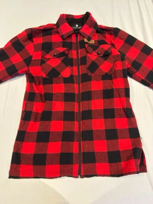 Röd och svart overshirt från Woodline - Snygg röd och svart overshirt från Woodline i klassiskt rutigt mönster. Skjortjackan har dragkedja framtill, två bröstfickor med knappar och mjukt tyg som känns skönt mot huden. Perfekt lager på lager-plagg för kyligare dagar.