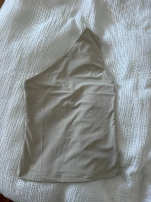 Beige oneshoulder topp - Snygg beige topp med oneshoulder-design. Toppen är ärmlös och har en stilren, minimalistisk look. Perfekt att matcha med jeans eller kjol för en trendig outfit. Använd en gång