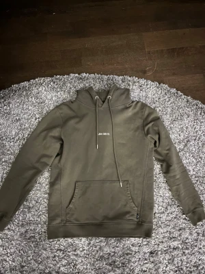 Olivgrön hoodie från Les Deux - Snygg olivgrön hoodie från Les Deux med broderad logga på bröstet. Klassisk känguruficka framtill, justerbar huva med dragsko och ribbade muddar. Perfekt för en avslappnad och trendig stil.