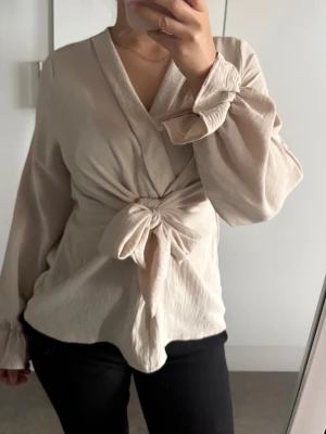 Beige omlottblus med knyt framtill - Trendig beige omlottblus med knyt i midjan och långa, vida ärmar med lätt volang vid ärmslut. Blusen har en djup V-ringning och är tillverkad i ett mjukt, lätt material som ger en avslappnad men stilren look.