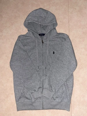 Ralph Lauren Zip Hoodie Grå – Helt Ny med Tags – Medium - Säljer en helt ny grå Ralph Lauren zip hoodie i storlek Medium. Tröjan är oanvänd. Tillverkad i mjukt och bekvämt material med klassisk passform, dragkedja framtill, huva med dragsko och den ikoniska broderade Ralph Lauren-loggan på bröstet.  Skick: Helt ny med tags Storlek: Medium (M) Färg: Grå Märke: Ralph Lauren  Perfekt för både vardag och en stilren, avslappnad look. Skicka gärna ett meddelande om du har frågor eller vill ha fler bilder.