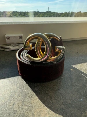 Vinrött mockabälte från Gucci - Snyggt vinrött bälte från Gucci i mocka med ikoniskt GG-spänne i guld. Bältet har en bred design och ger en lyxig känsla till din outfit. Perfekt accessoar för att lyfta vilken look som helst.
