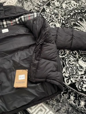 Svart pufferjacka från Burberry - Snygg svart pufferjacka från Burberry med klassiskt rutigt foder på insidan och tydlig logga på ärmen. Jackan har en vadderad design, hög krage och dragkedja. Perfekt för kalla dagar när du vill hålla stilen.