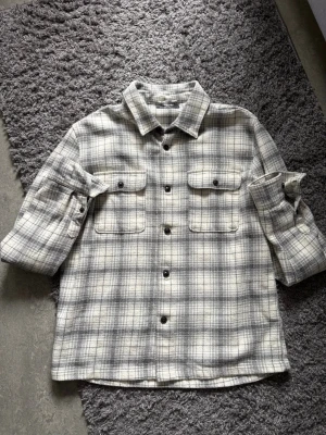 Overshirt dressmann - Snygg overshirt i gråvitt rutigt mönster med två bröstfickor och svarta knappar. Skjortan har klassisk krage och långa ärmar med knapp vid manschetten. Perfekt lager på lager-plagg i mjukt tyg.