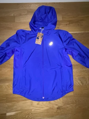 Lila vindjacka från Asics med huva - Snygg lila vindjacka från Asics med huva och hel dragkedja framtill. Jackan har reflekterande Asics-logga på bröstet och är tillverkad i lätt, vindtätt material. Perfekt för löpning eller utomhusaktiviteter när det blåser.