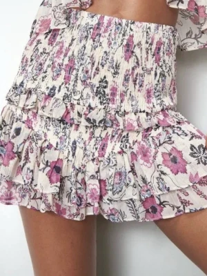 Blommig volangkjol i ljusa toner - Supersöt kortkjol med blommigt mönster i lila, rosa och svart på ljus botten. Kjolen har smock i midjan och flera volanger som ger en flowy känsla. 