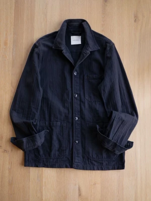 Svart overshirt från A Day's March - Stilren svart overshirt från A Day's March med tre stora utanpåliggande fickor och knäppning framtill. Overshirten har klassisk krage och är tillverkad i ett kraftigare bomullstyg som ger en snygg och avslappnad look.