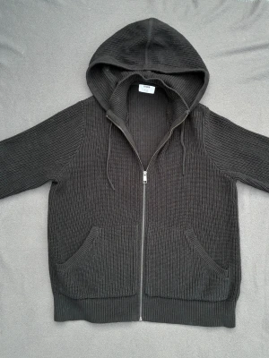 Svart ribbstickad hoodie - Riktigt clean ribbstickad hoodie. Storlek M, för stor för han på bilden(rekommenderad längd 180-192). Fint skick, ljuset gör att den ser lite missfärgad ut men färgen är som på bild 4.