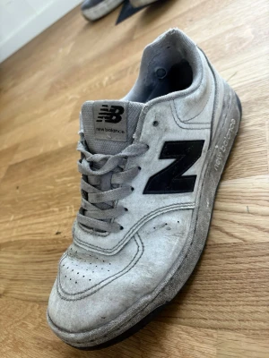New Balance sneakers - Säljer ett par vita New Balance sneakers. Skorna har lågt skaft, vit snörning och en robust sula. Materialet är mocka och textil, och designen är klassisk med perforerade detaljer framtill.