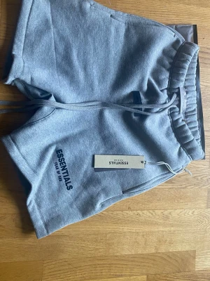Grå shorts från Essentials XXS - Gråa shorts från Essentials med elastisk midja och snörning. Mjukt material i bomull och polyester, perfekt för en avslappnad stil. Logga tryckt på vänster ben. Snygg och enkel design som passar till många outfits.
