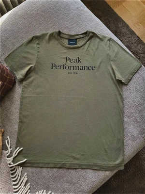 Olivgrön Peak Performance t-shirt - Snygg olivgrön t-shirt från Peak Performance med svart tryck på bröstet. Klassisk rund halsringning och korta ärmar. Tillverkad i mjuk bomull som känns skön mot huden. Perfekt för en avslappnad och stilren look. Storlek 160 cm, 14 år. 