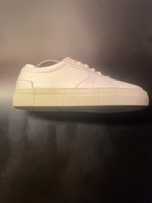 Vita sneakers från Axel Arigato - Säljer ett par stilrena vita sneakers från Axel Arigato. Skorna är låga med klassisk siluett, tillverkade i slätt läder och har en tjock vit sula. Diskret logga på sidan och ton-i-ton snörning. Perfekta för en clean och modern look.