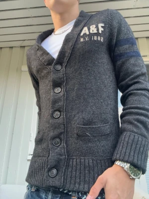 Vintage Abercrombie and Fitch Kofta Cardigan - En riktigt snygg grå kofta från det trendiga Abercrombie and Fitch🤩 En tröja som verkligen passar perfekt för att gå med stil nu när hösten närmar sig🍁 Tygets tjocklek och kvalitet ljuger inte, denna kommer hålla din kropp sällskap i många år🙌 Stl M men passar snarare som S/M.  Längd: 65cm   Bredd mellan armhålor: 48 cm.      Jag är 176cm lång och på mig passar den perfekt - se bild🤩