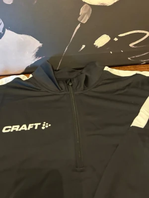 Svart långärmad träningströja Craft - Svart långärmad träningströja från Craft med vit logga på bröstet och vita detaljer längs axlarna. Tröjan har halv dragkedja framtill och är tillverkad i ett lätt och funktionellt material som passar perfekt för träning.