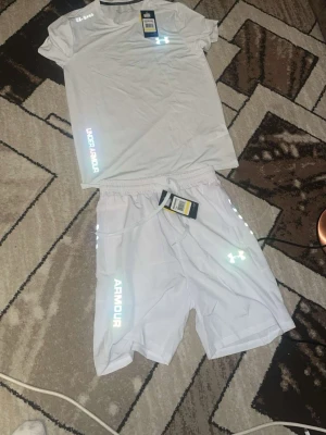 Vitt träningsset från Under Armour - Säljer ett fräscht vitt träningsset från Under Armour med t-shirt och shorts. Setet har reflekterande loggor och text, samt elastisk midja på shortsen. Perfekt för gymmet eller löpning. Materialet är lätt och andas bra.