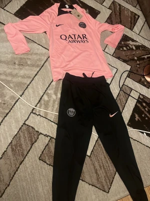 PSG träningsset rosa/svart Nike - Säljer ett PSG träningsset från Nike med långärmad rosa tröja och svarta byxor. Tröjan har PSG-logga och Qatar Airways-tryck framtill, medan byxorna har PSG-emblem och rosa Nike-logga. Perfekt för fotbollsträning eller chill.