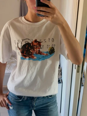 Vit t-shirt med kattmotiv - Säljer en vit t-shirt med ett färgglatt tryck av en katt och text på framsidan, alltså från Masion Martin Marigela. Med prislappen på. T-shirten har rund hals med dubbla stygn och korta ärmar. Passformen är normal och materialet känns mjukt och bekvämt. Storlek S,  100% bomull