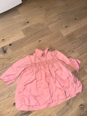 Rosa klänning med volangkrage - Söt rosa klänning med volangkrage och smockdetalj i midjan. Klänningen har lång ärm med volangkant och är gjord i ett mjukt tyg, perfekt för en trendig och bekväm look.Rosa klänning med volangkrage