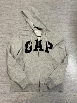 Grå hoodie med dragkedja från GAP - Snygg grå hoodie från GAP med dragkedja framtill och stor svart logga på bröstet. Tröjan har huva med vita snören och ribbade muddar vid ärmslut och nederkant. Perfekt för en avslappnad och trendig look.