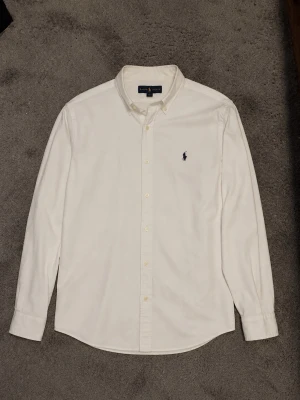 Polo Ralph Lauren Skjorta - Klassisk vit skjorta från Ralph Lauren med button-down krage och broderad logga på bröstet. Skjortan har långa ärmar och knäppning framtill. Perfekt till en clean och stilren look.