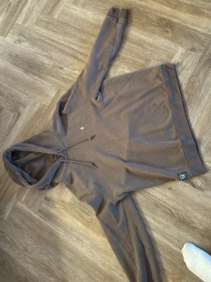 Brun hoodie med liten logga Morris  - Säljer en brun hoodie med huva och dragsko. Tröjan har en liten broderad logga på bröstet och en patch nertill vid midjan. Tillverkad i mjukt bomullsmaterial och har ribbade muddar vid ärmslut och nederkant. Den kan även passa L