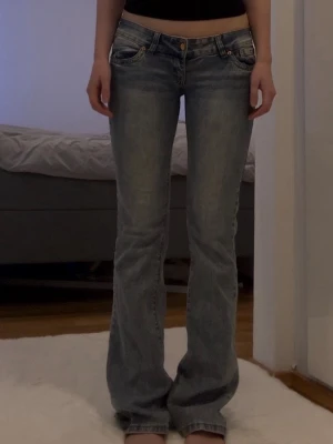 Ljusblå bootcut jeans med låg midja - Säljer ett par ljusblå bootcut jeans med låg midja och klassiska fem fickor. Jeansen har en avslappnad passform och coola detaljer för en trendig look. Perfekta till en enkel topp eller hoodie. 84 cm innerbenslängd.
