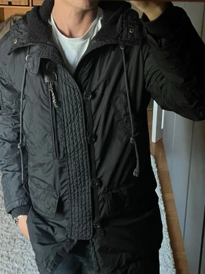 Peak Performance Detroit parka i svart - stl M - Peak Performance Detroit parka i svart, storlek M med skal i 100% polyamid, quiltat foder i polyester och avtagbar luva med pilefoder.  Använd med ett hål i fodret vid luvans fäste i nacken, ca 3 cm, där vadderingen syns igenom. Syns inte utifrån och påverkar inte funktionen, se bild 5. I övrigt hel och ren, inga fläckar.  Personen på bilden är 189cm, väger 78kg.  Rök- och djurfritt hem. Skriv gärna om du vill ha fler bilder eller undrar något!