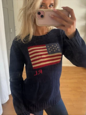 Äkta ralph Lauren - Mörkblå stickad tröja med en stor amerikansk flagga på bröstet och röda bokstäver 'RL' under flaggan. Tröjan har rund halsringning, långa ärmar och ribbstickade muddar. Sparsamt använd och enda defekten är det som syns i flaggan pga slitage