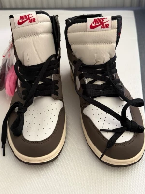 Nike Air Jordan 1 Travis Scott High - Säljer ett par Nike Air Jordan 1 Travis Scott High med bruna, vita och svarta färgblock. Skorna har ikonisk omvänd swoosh i svart, 'Cactus Jack'-detaljer och högt skaft. Tillverkade i läder och mocka, med svarta skosnören och extra snören i rosa och rött.