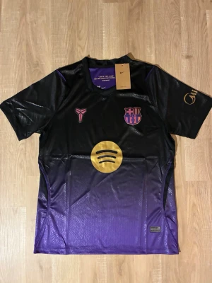 Svart-lila FC Barcelona matchtröja 26/27 - Snygg FC Barcelona matchtröja i svart och lila med guldfärgade detaljer och loggor. Tröjan har korta ärmar, rund hals och är tillverkad i ett lätt, ventilerande material. Perfekt för fotbollsträning eller att bära som supporter. HELT NY!