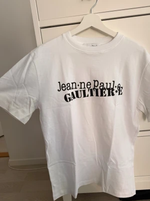 Vit t-shirt Jean Paul Gaultier - Snygg vit t-shirt från Jean Paul Gaultier med svart tryck framtill. Klassisk rund hals och korta ärmar. Perfekt till jeans eller shorts för en clean och trendig look.