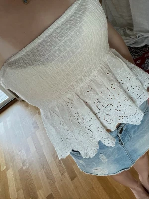 Vit axelbandslös spets tubtopp - Söt vit axelbandslös tubtopp med smockad överdel och volangkant i broderad spets. Toppen har en luftig och somrig känsla med fina detaljer i spetsen. Perfekt att styla till kjol eller jeans för en fräsch look.
