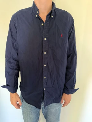 Mörkblå skjorta från Polo Ralph Lauren - Klassisk mörkblå skjorta från Polo Ralph Lauren med knappkrage och röd broderad logga på bröstet. Skjortan har långa ärmar och vita knappar framtill. Hör av dig om du har några frågor eller vill ha fler bilder. Pris kan diskuteras! 