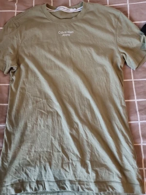 Olivgrön t-shirt från Calvin Klein Jeans - Säljer en olivgrön t-shirt från Calvin Klein Jeans med vit logga tryckt på bröstet. Klassisk rund hals och korta ärmar. Tillverkad i mjuk bomull för skön känsla och enkel stil.
