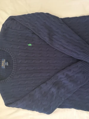 Mörkblå kabelstickad tröja Polo Ralph Lauren - Kabelstickad mörkblå tröja från Polo Ralph Lauren med rund halsringning och den klassiska gröna logotypen broderad på bröstet. Tröjan har långa ärmar och ribbade muddar vid ärmslut och nederkant. Perfekt för en stilren look.