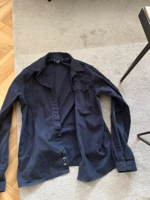 Svart overshirt med knappar - Stilren svart overshirt med klassisk krage och knäppning framtill. Skjortan har en bröstficka med lock och långa ärmar med knapp vid manschetten. Perfekt att bära öppen över en t-shirt för en avslappnad look.