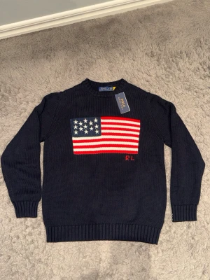 Mörkblå stickad tröja Polo Ralph Lauren - Mörkblå stickad tröja från Polo Ralph Lauren med amerikansk flagga i rött, vitt och blått på bröstet samt broderade initialer 'RL' i rött. Tröjan har rund halsringning och ribbade muddar vid ärmslut och nederkant. Perfekt för dig som gillar klassisk och preppy stil.