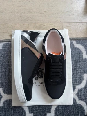 Burberry svart/vit låg sneaker - Snygga sneakers från Burberry i svart med klassiskt rutmönster i vitt och beige på sidan. Skorna har vita sulor, svarta skosnören och en stilren design i textil och läder. Perfekta för dig som vill ha en trendig och tidlös look.