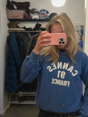 Blå sweatshirt med tryck - Blå sweatshirt med texten 'CANNES 91 FRANCE' i vitt tryck på framsidan. Tröjan har rund halsringning och avslappnad passform, perfekt för en chill och sportig look.