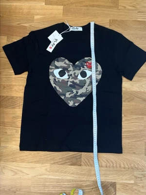 Svart Comme des Garçons PLAY t-shirt - Svart t-shirt från Comme des Garçons PLAY med stort hjärta i camouflage-mönster och ett litet rött hjärta med ögon på bröstet. Klassisk rund hals och korta ärmar. Perfekt för dig som gillar streetwear och coola prints.