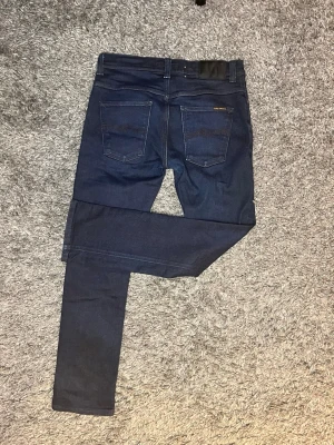 Mörkblå slimfit nudie jeans - Snygga mörkblå slimfit jeans från Nudie med klassisk femficksdesign och diskreta sömmar. Jeansen har en smal passform och är tillverkade i stretchigt denimtyg för extra komfort. Perfekta för en trendig och stilren look. Jeansen är i strl w32 l32. Skriv vid fler frågor🙌