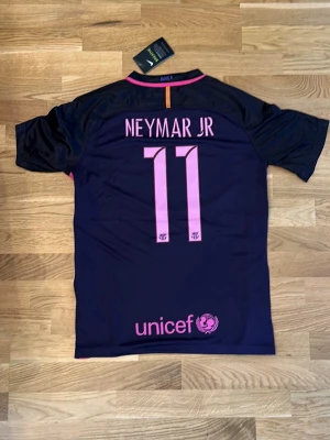 Barcelona Neymar Jr matchtröja lilla 2014/2015 - Säljer en mörklila Barcelona matchtröja från Nike med Neymar Jr och nummer 11 på ryggen. Tröjan har rosa detaljer, klubbmärke, Champions League-märke på ärmen och Qatar Airways som sponsor framtill. Perfekt för dig som älskar fotboll och Barca!