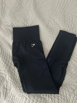 Svarta leggings med hög midja - Snygga svarta leggings från Gymshark med hög ribbad midja och diskret logga på höften. Stretchigt material som sitter tight och formar sig efter kroppen. Perfekta för träning eller chill dagar.