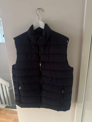 Svart quiltad dunväst med dragkedja - Stilren svart dunväst med hög krage och quiltat mönster. Västen har två fickor med dragkedja framtill och stängs med en hel dragkedja. Perfekt att slänga över en hoodie eller tröja för extra värme. Materialet är mjukt och puffigt för skön komfort.
