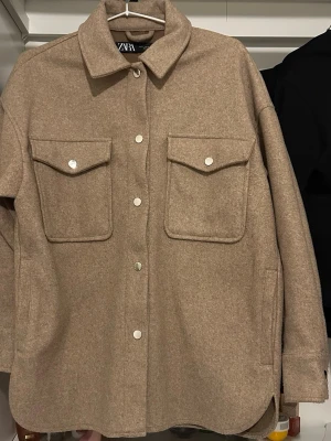 Beige/brun  jacka från ZARA - Stilren beige/brun från ZARA  med klassisk krage och tryckknappar framtill. Två stora bröstfickor med lock och knapp, samt diskreta sidofickor. Rak passform och långa ärmar med knapp vid ärmslut. Perfekt lager på lager-plagg.