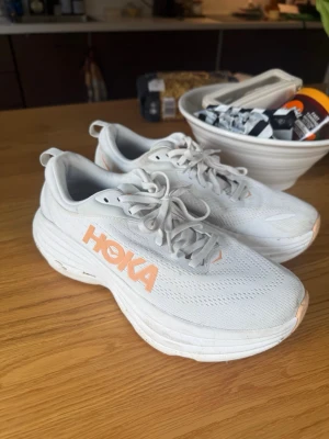Vita Hoka skor - Säljer ett par vita Hoka sneakers med orange logga på sidan. Modell: Bondi 8.  Skorna har en chunky sula och ovandel i mesh för bra ventilation. Klassisk låg modell med snörning och detaljer i vitt och ljusorange. Perfekta för dig som gillar sportig och clean stil. Storlek 41, 1/3 men är väldogt små i storlek och passar mig som har 39-40 vanligtvis. Använd max 5 gånger
