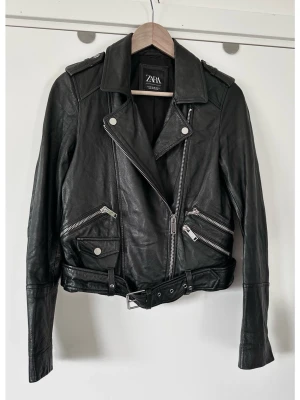 ZARA Black Leather Biker Jacket - size xs - Stilren cool svart skinnjacka i bikerstil från ZARA i storlek XS med silverfärgade dragkedjor och knappar. Klassisk modell med asymmetrisk dragkedja framtill, slag med tryckknappar och axelklaffar. Det finns flera fickor med dragkedja fram samt en liten ficka med lock och tryckknapp. Ärmsluten har dragkedjor och jackan har ett justerbart bälte i midjan. 100% äkta läder. En tidlös klassiker som kommer hålla säsong efter säsong. Nypris 2600:-. Mycket gott skick!
