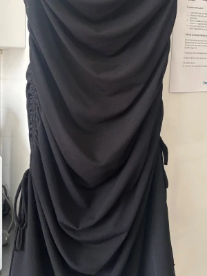 Svart draperad kortklänning - Säljer en svart kortklänning med snygga draperingar och snörningsdetaljer på sidorna. Klänningen har en figurnära passform och är perfekt för dig som gillar en stilren och trendig look.